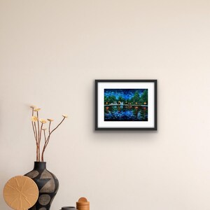 May include: A framed painting depicts a nighttime waterscape with a bridge, trees, and reflections in the water. The artwork features a dark blue sky with stars and colorful lights. The painting is in a black frame.