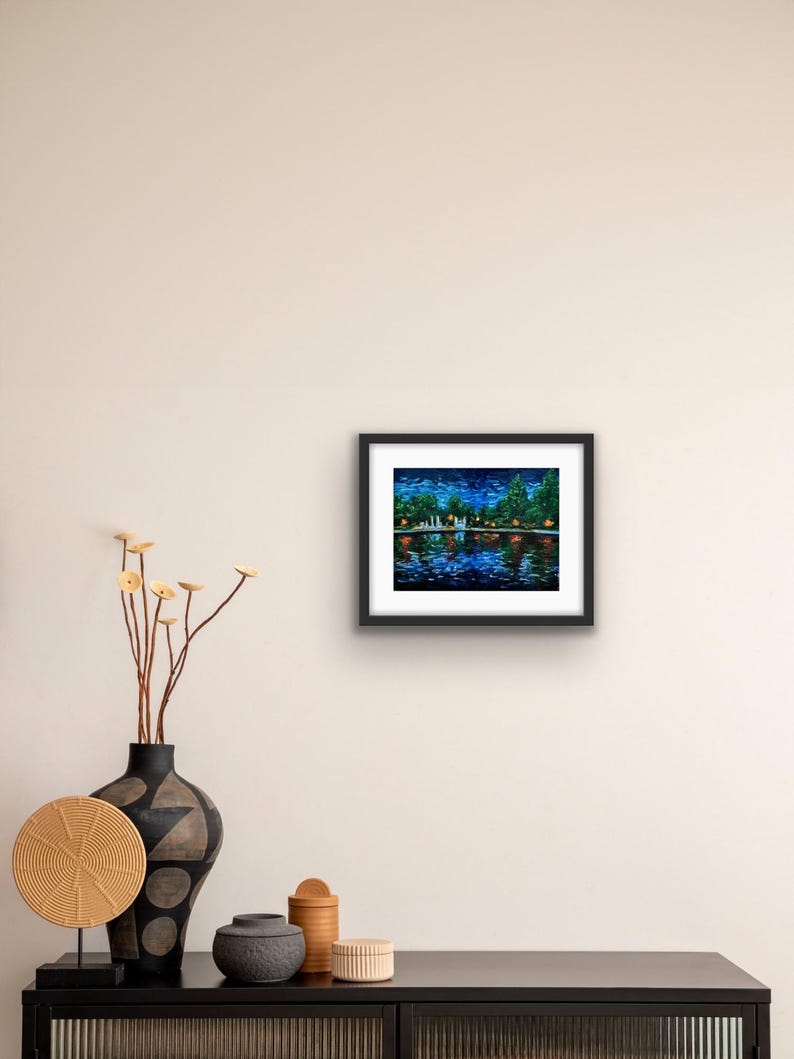 May include: A framed painting depicts a nighttime waterscape with a bridge, trees, and reflections in the water. The artwork features a dark blue sky with stars and colorful lights. The painting is in a black frame.