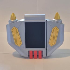 May include: A silver and gold-colored device with a black screen. The device has gold accents, including two conical shapes on top and two diamond-shaped designs on the sides. Three red rectangular shapes are at the bottom.