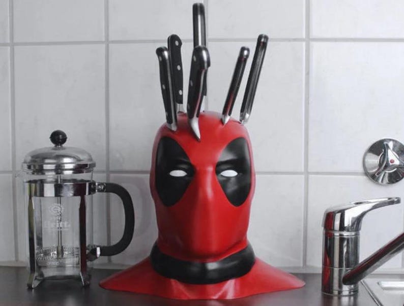 May include: A red and black Deadpool-themed knife block holding several black-handled knives. The knife block is shaped like Deadpool's mask. A stainless steel French press and a faucet are also visible.