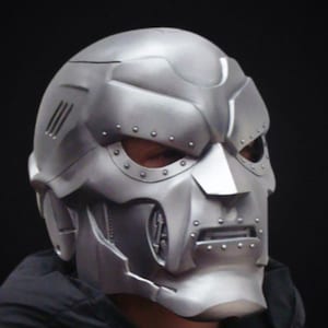 May include: A silver metal helmet with a detailed design, including a raised ridge and a series of small rivets. The helmet has a visor and a chin guard.