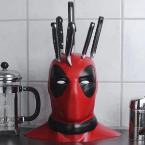 May include: A red and black Deadpool-themed knife block holding several black-handled knives. The knife block is shaped like Deadpool's mask. A stainless steel French press and a chrome faucet are also visible.