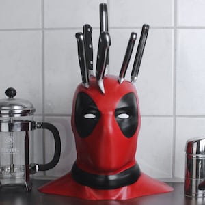 May include: A red Deadpool-themed knife block with black detailing, holding kitchen knives. The knife block is shaped like Deadpool's mask. A French press coffee maker and a chrome tap are also visible.