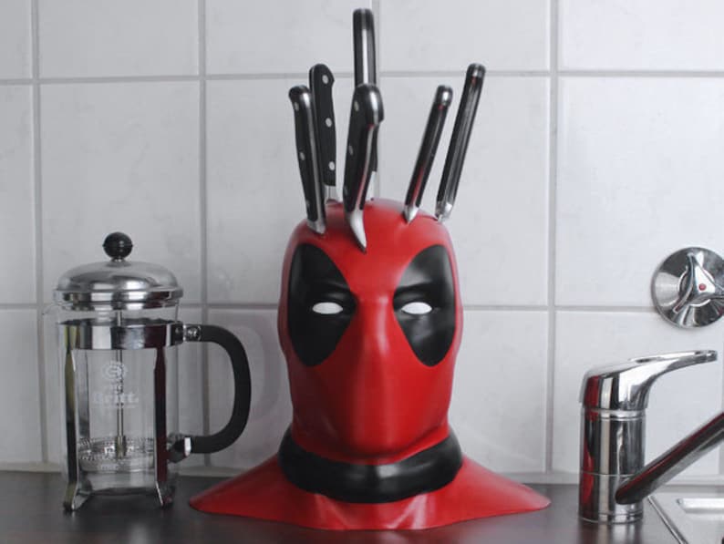 May include: A red Deadpool-themed knife block with black accents, holding several kitchen knives. The knife block is shaped like Deadpool's mask. A French press coffee maker and a chrome faucet are also visible.