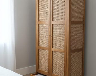 May include: Wooden wardrobe with woven cane panels and two drawers. The wardrobe is made of light-colored wood and has a natural finish. A vase with greenery sits on top. The drawers are partially open, revealing folded linens.