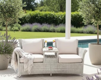 May include: A light gray wicker outdoor loveseat with a small side table, two cream-colored cushions, and a matching throw blanket. Two olive trees in large pots flank the loveseat. Two drinks with fruit are on the table.