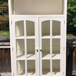 May include: White wooden cabinet with glass doors and shelves. The cabinet has a curved top and a single drawer at the bottom.
