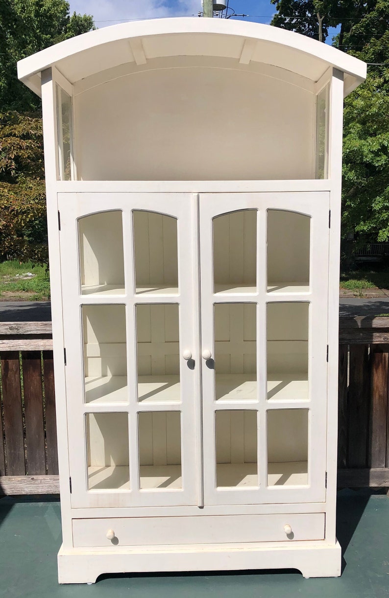 May include: White wooden cabinet with glass doors and shelves. The cabinet has a curved top and a single drawer at the bottom.