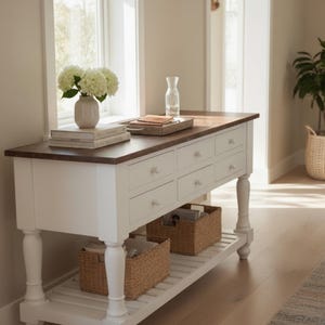 May include: A white and brown console table with six drawers and two wicker baskets on a lower shelf. The table has a dark brown top and white legs. A vase of white flowers and books sit on top.