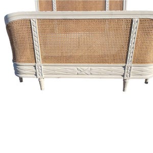 May include: A white painted bed frame with a woven cane headboard and footboard. The frame has decorative carved details and four legs. The headboard is tall and rectangular, while the footboard is shorter and curved.