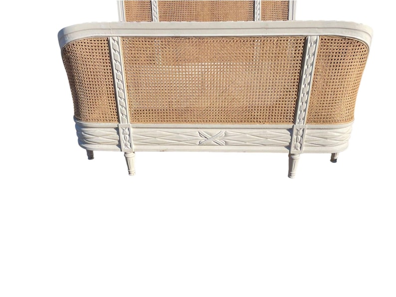 May include: A white painted bed frame with a woven cane headboard and footboard. The frame has decorative carved details and four legs. The headboard is tall and rectangular, while the footboard is shorter and curved.