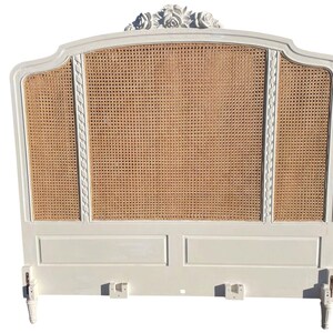May include: A white, ornate headboard with a woven cane centre panel. The headboard features a curved top with floral carvings and decorative trim. The frame is painted white, and the cane panel has a natural, light brown colour. The headboard has two legs.