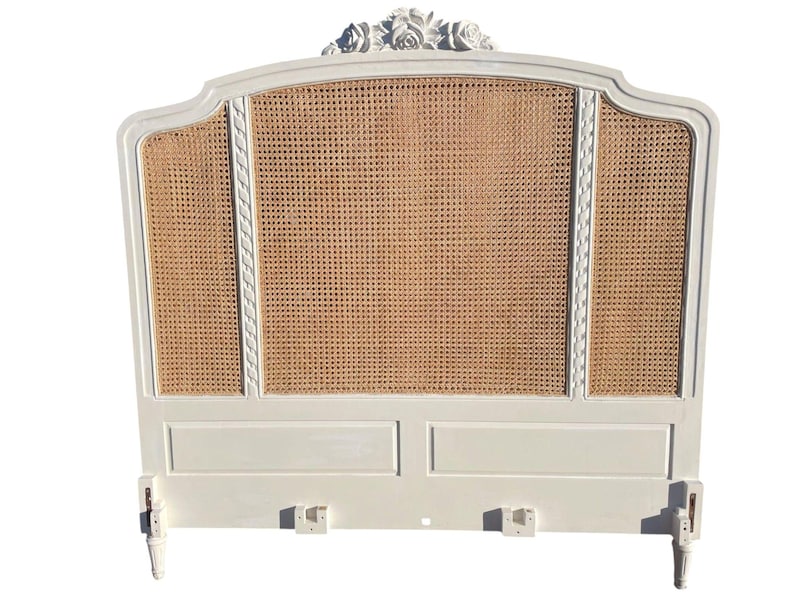 May include: A white, ornate headboard with a woven cane centre panel. The headboard features a curved top with floral carvings and decorative trim. The frame is painted white, and the cane panel has a natural, light brown colour. The headboard has two legs.