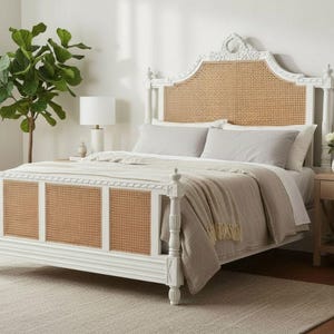 May include: A white bed frame with a woven rattan headboard and footboard panels. The bed is made up with light gray bedding and a beige throw blanket. A matching nightstand with a lamp and a plant are next to the bed.