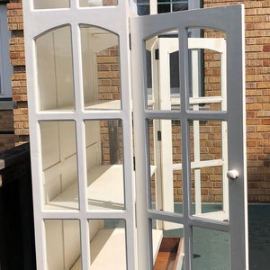 May include: White wooden cabinet with glass doors and shelves. The cabinet has a drawer on the bottom and is open with the door ajar.