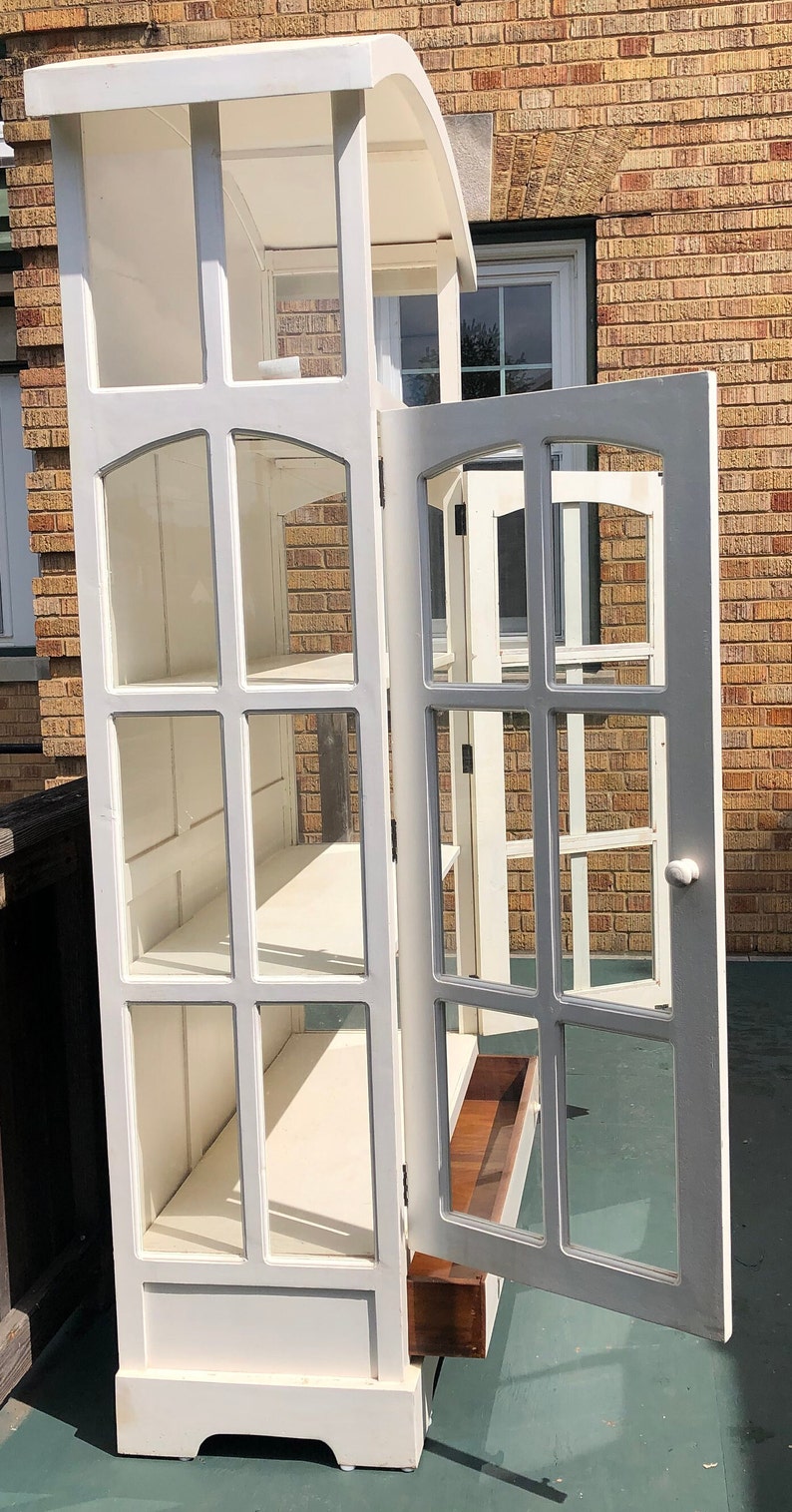 May include: White wooden cabinet with glass doors and shelves. The cabinet has a drawer on the bottom and is open with the door ajar.
