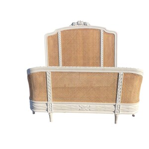 May include: A white and natural-toned bed frame with a woven cane headboard and footboard. The frame has decorative carvings and a curved design, with a classic, elegant aesthetic. The bed frame is a double or queen size.