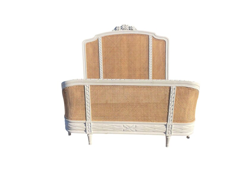 May include: A white and natural-toned bed frame with a woven cane headboard and footboard. The frame has decorative carvings and a curved design, with a classic, elegant aesthetic. The bed frame is a double or queen size.