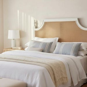 May include: A bedroom scene featuring a white bed with a woven headboard and light blue striped pillows. A beige throw blanket with fringe is draped across the bed. Two white lamps sit on nightstands on either side of the bed.