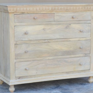 Puede incluir: Una c&oacute;moda de madera clara con cinco cajones y una moldura decorativa alrededor de la parte superior. La c&oacute;moda tiene patas torneadas y tiradores de pomo simples.