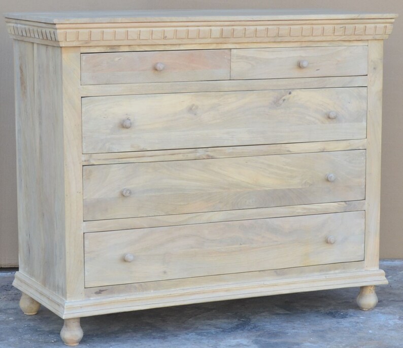 Puede incluir: Una c&oacute;moda de madera clara con cinco cajones y una moldura decorativa alrededor de la parte superior. La c&oacute;moda tiene patas torneadas y tiradores de pomo simples.
