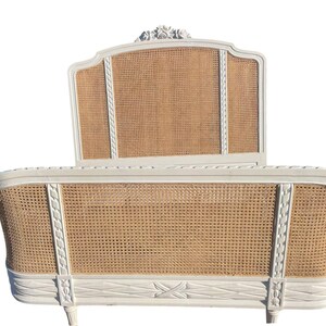 May include: A white, vintage-style bed frame with a woven cane headboard and footboard. The frame has decorative carvings and a curved design. The headboard features a floral accent at the top. The bed frame is a classic piece of bedroom furniture.
