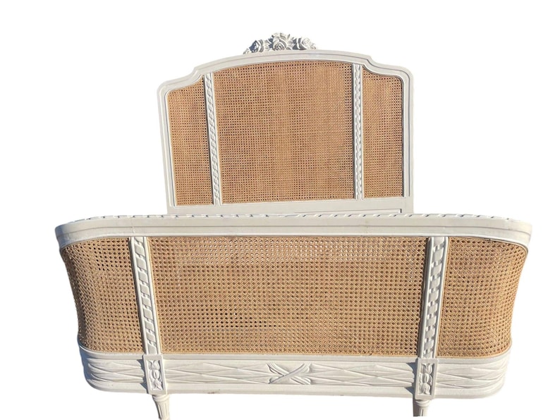 May include: A white, vintage-style bed frame with a woven cane headboard and footboard. The frame has decorative carvings and a curved design. The headboard features a floral accent at the top. The bed frame is a classic piece of bedroom furniture.