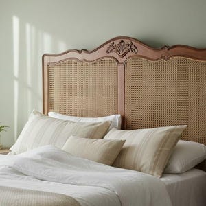 May include: A wooden headboard featuring a natural cane webbing insert and a carved floral detail at the center top. The headboard has a warm brown finish and a curved silhouette, positioned behind a bed with white and beige striped pillows.