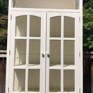 May include: White wooden cabinet with glass doors and a single drawer. The cabinet has a curved top and is decorated with a simple design.