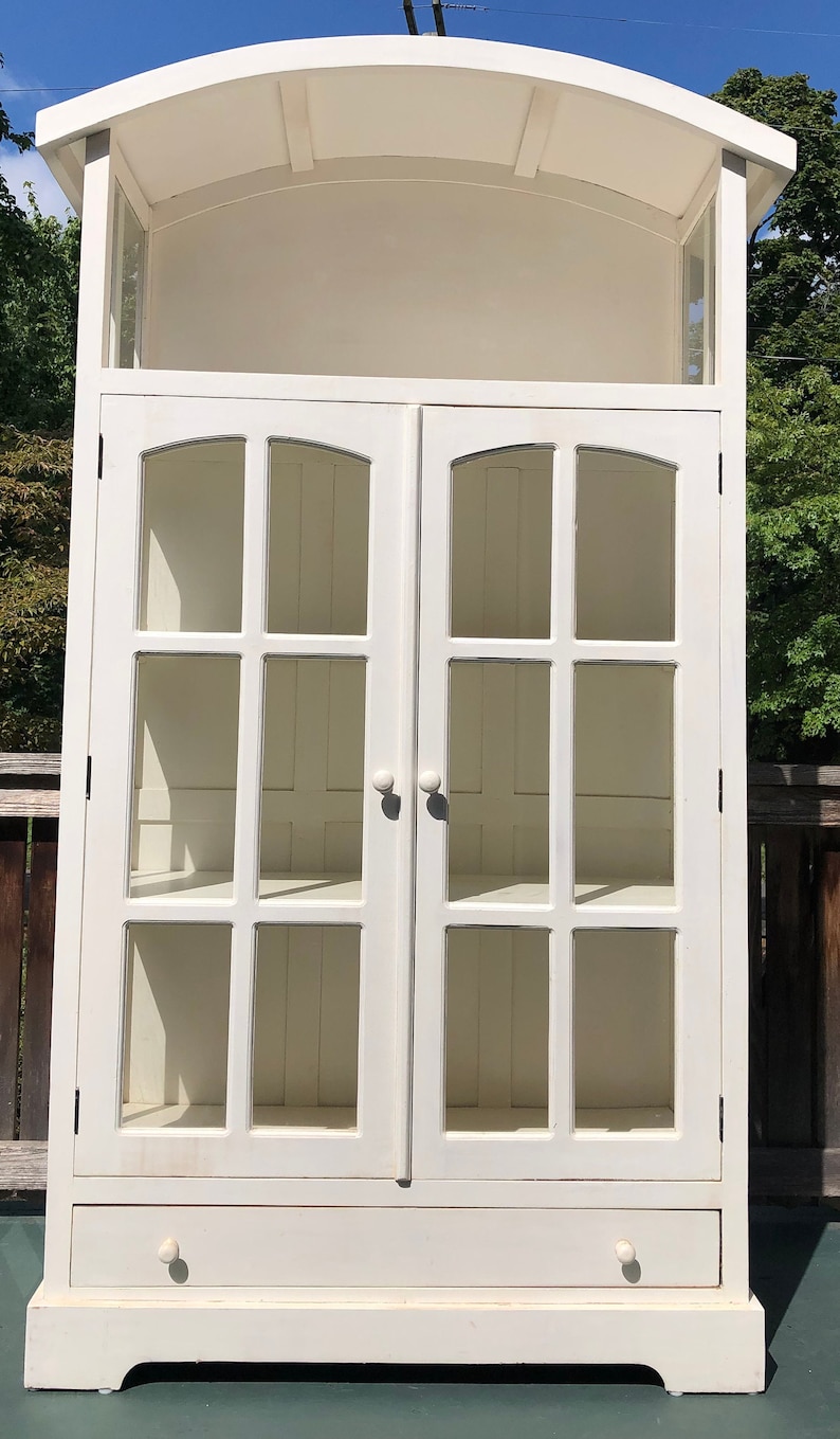 May include: White wooden cabinet with glass doors and a single drawer. The cabinet has a curved top and is decorated with a simple design.