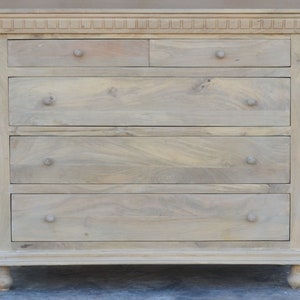 Puede incluir: Una c&oacute;moda de madera clara con cinco cajones y una parte superior decorativa. La c&oacute;moda tiene patas torneadas y un dise&ntilde;o sencillo.