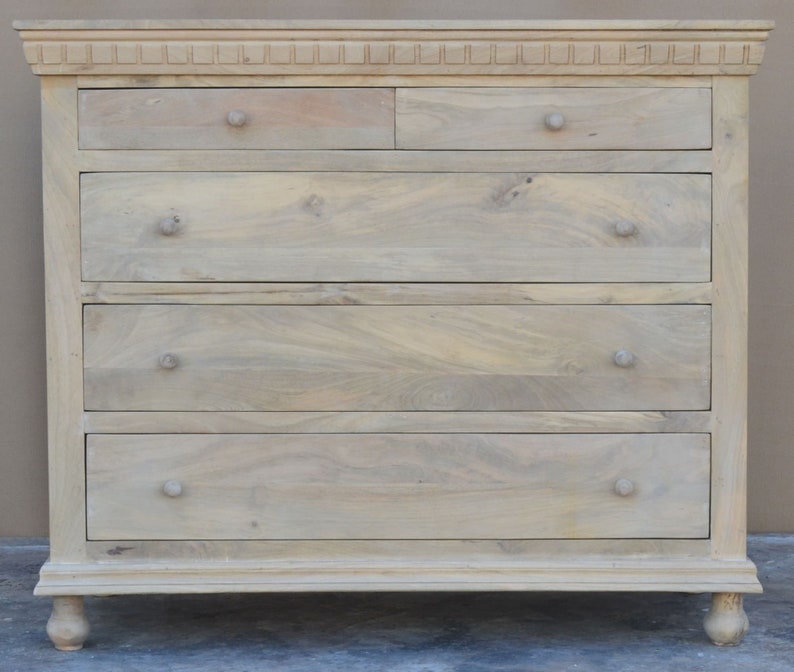Puede incluir: Una c&oacute;moda de madera clara con cinco cajones y una parte superior decorativa. La c&oacute;moda tiene patas torneadas y un dise&ntilde;o sencillo.