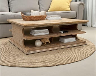 May include: A light-coloured wooden coffee table with two shelves, a woven basket, and books. The table sits on a round, woven jute rug. A grey sofa with a yellow cushion is in the background.