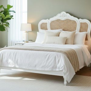 May include: A white bed frame featuring a curved, ornate headboard with natural woven cane paneling. The bed is dressed in white linens, including a textured beige throw blanket at the foot, and is flanked by two matching bedside tables with lamps.