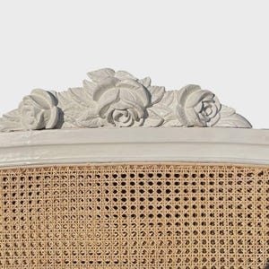 May include: A close-up of a white headboard with a woven cane panel. The top features a decorative floral carving of three roses and leaves. The headboard is a light neutral colour, suitable for a bedroom.