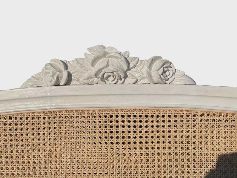 May include: A close-up of a white headboard with a woven cane panel. The top features a decorative floral carving of three roses and leaves. The headboard is a light neutral colour, suitable for a bedroom.
