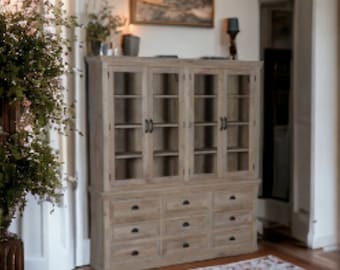 May include: A wooden cabinet with glass doors and drawers. The cabinet has a light wood finish and is decorated with black metal hardware. The cabinet is in a room with a rug on the floor.