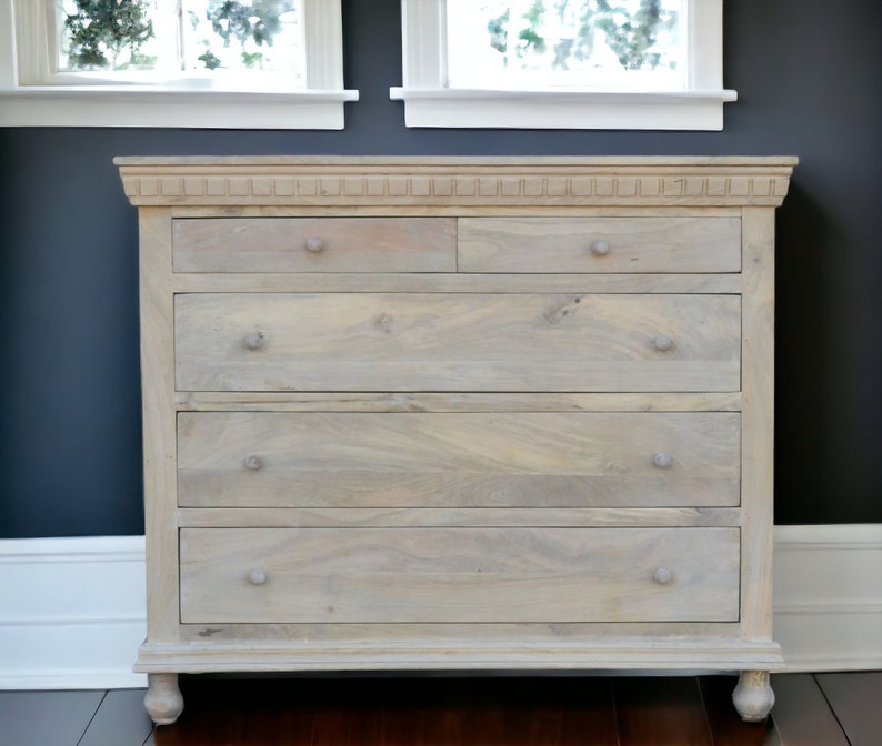 Puede incluir: Una c&oacute;moda de madera gris claro con cinco cajones y tiradores simples. La c&oacute;moda tiene un acabado ligeramente desgastado y se encuentra sobre un suelo de madera oscura.