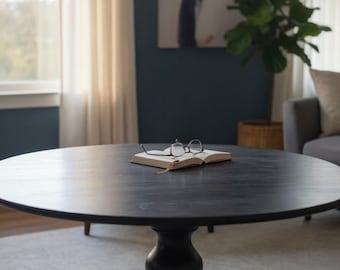 May include: A dark, round wooden coffee table with a pedestal base. An open book and eyeglasses rest on the table's surface. The table is in a living room setting with a painting, plant, and window.