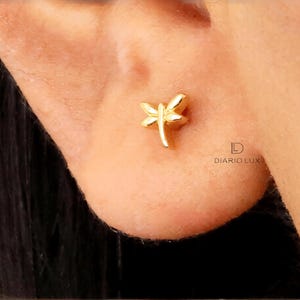 May include: A small gold-toned stud earring shaped like a dragonfly with delicate wings and a slender body. The earring is positioned on an earlobe. The text DIARIO LUX is printed in black near the earring, featuring a stylized letter D logo.