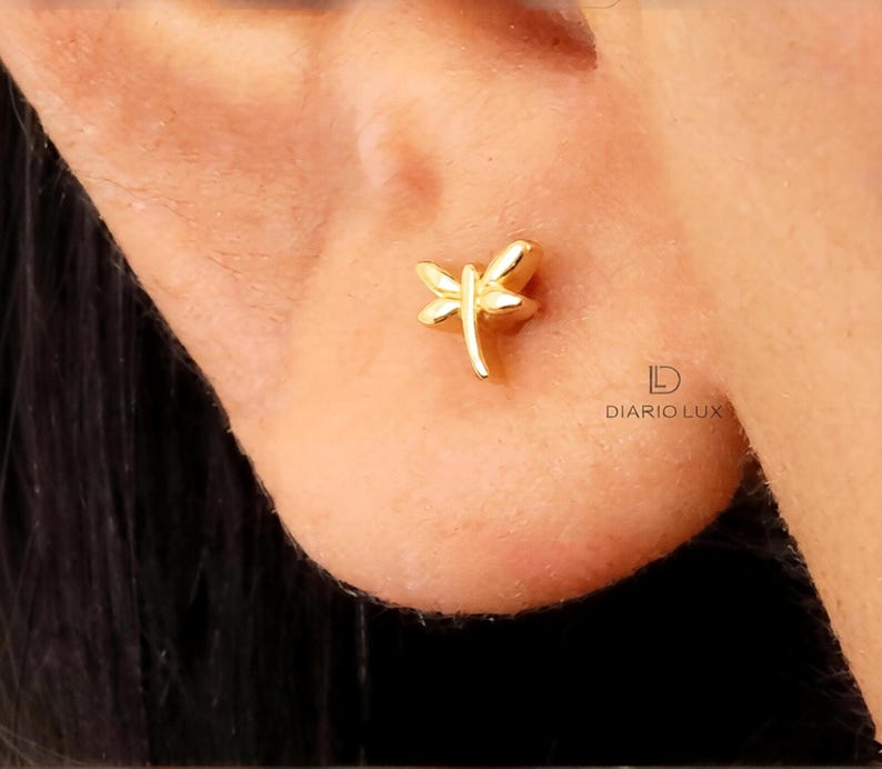 May include: A small gold-toned stud earring shaped like a dragonfly with delicate wings and a slender body. The earring is positioned on an earlobe. The text DIARIO LUX is printed in black near the earring, featuring a stylized letter D logo.