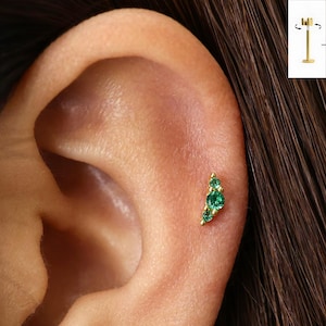 May include: A gold flat back stud earring featuring a curved cluster of three round green gemstones. The jewellery piece includes a thin metal post with a circular base, shown in an inset diagram, designed for secure placement in the ear cartilage.