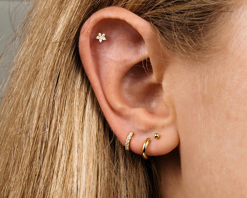 May include: A close-up of a person's ear with three gold earrings. The top earring is a small flower with clear stones. The middle earring is a small hoop with clear stones. The bottom earring is a small hoop with a ball.