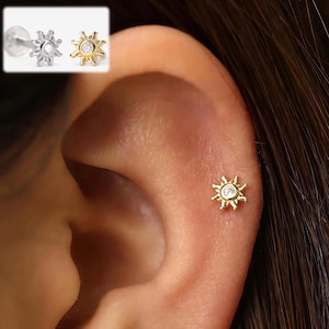 Puede incluir: Un pendiente dorado en forma de sol con una piedra central transparente se muestra en una oreja. Un segundo pendiente, en plata y oro, se muestra en la esquina superior izquierda. El pendiente es un pendiente de botón.