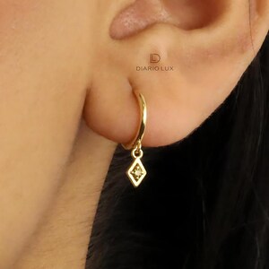 Pode incluir: Brincos de argola de ouro com um pequeno pingente em forma de diamante pendurado na parte inferior. O pingente tem um pequeno diamante &uacute;nico no centro.