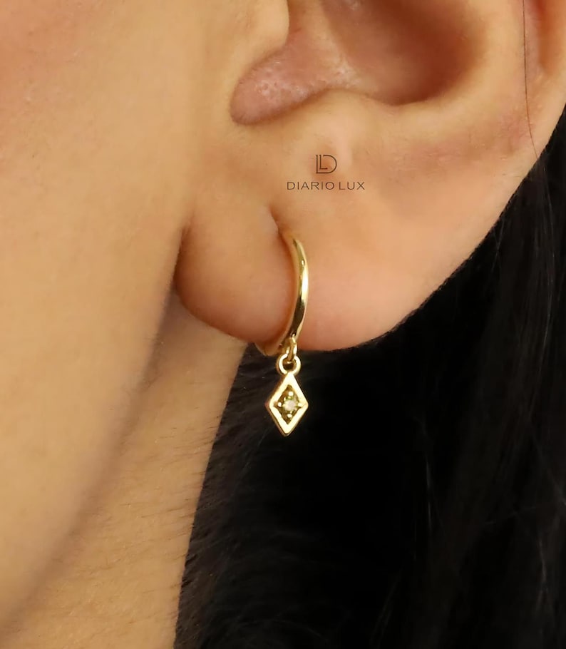 Pode incluir: Brincos de argola de ouro com um pequeno pingente em forma de diamante pendurado na parte inferior. O pingente tem um pequeno diamante &uacute;nico no centro.