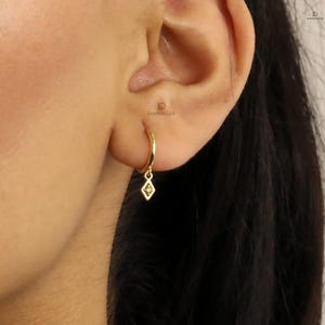 Pode incluir: Brincos de argola de ouro com um pequeno pingente em forma de diamante pendurado em cada argola.