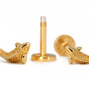 May include: A pair of gold-colored fish-shaped body jewelry pieces. The jewelry is designed to be worn as a cartilage piercing. The fish are detailed with scales and have a realistic appearance.