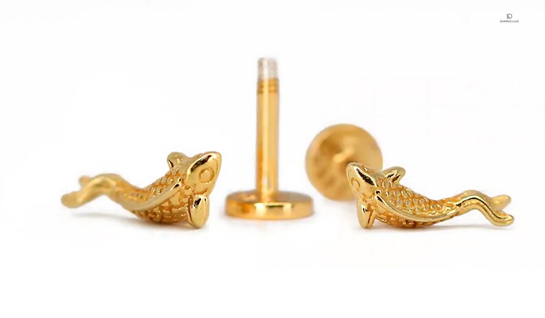 May include: A pair of gold-colored fish-shaped body jewelry pieces. The jewelry is designed to be worn as a cartilage piercing. The fish are detailed with scales and have a realistic appearance.