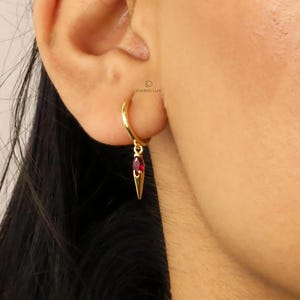 May include: Gold hoop earrings with a dangling red gemstone and a pointed gold charm.